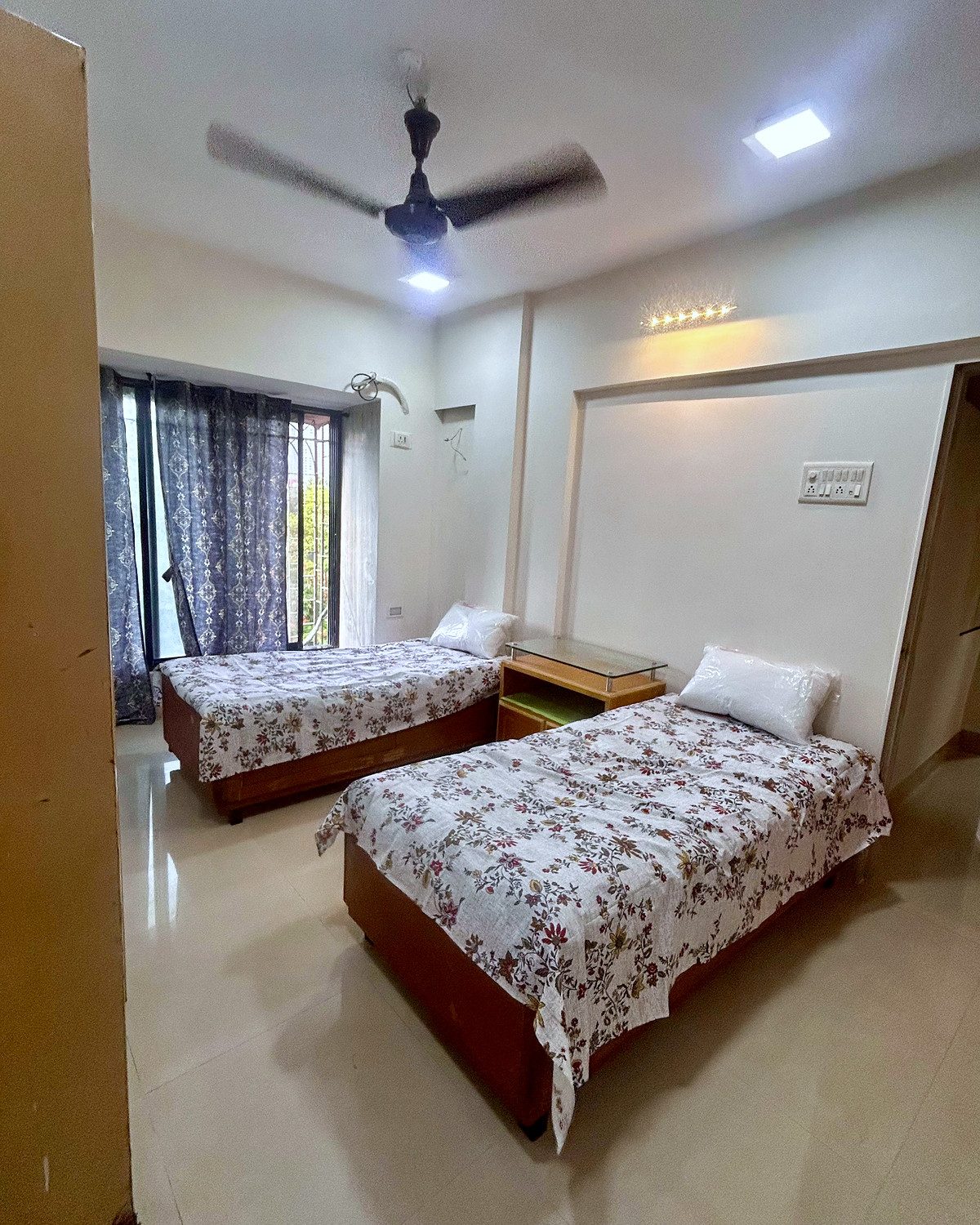 A sharing room with two beds set well apart, a large curtained window and daylight across the floor