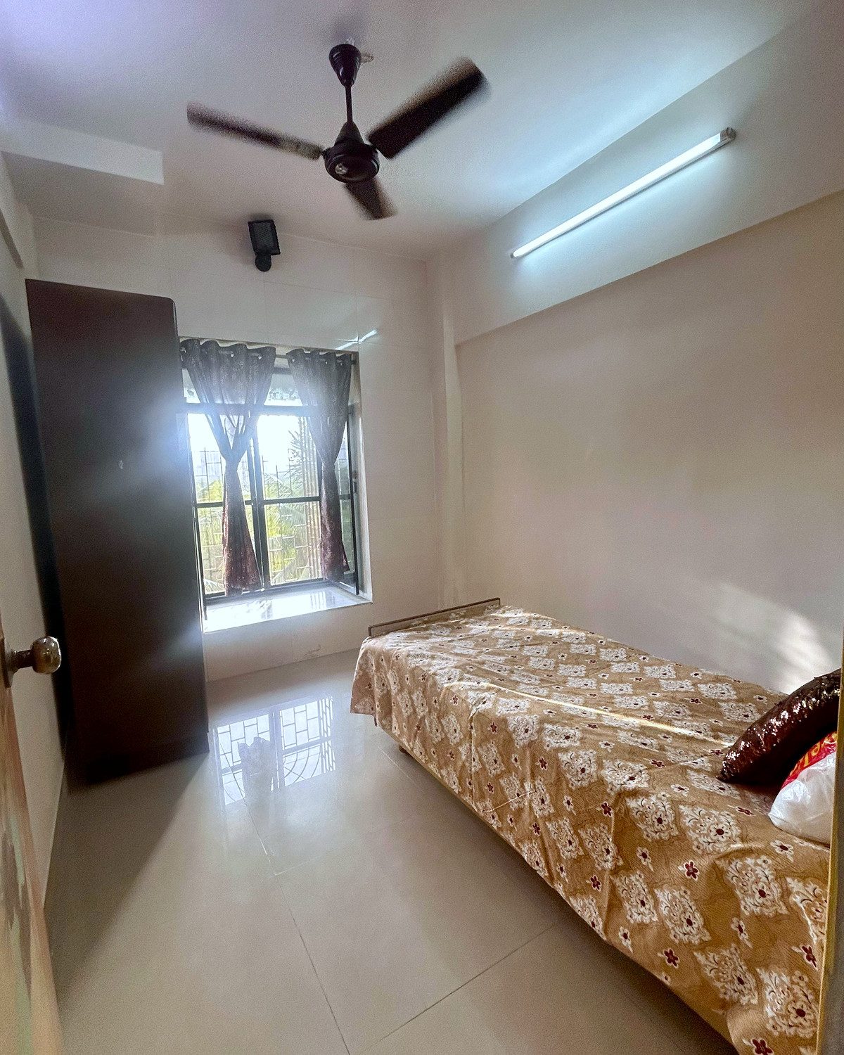 A single room lit by a tall window with clear floor space beside the bed